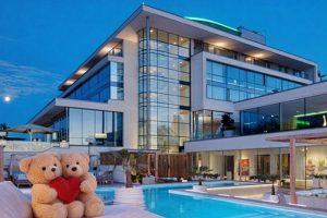 Två nallar med ett rött hjärta framför ett modernt hotell med pool under kvällshimlen.