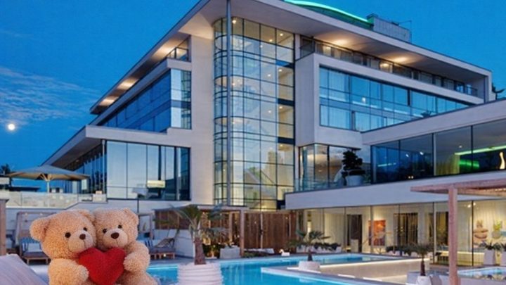 Två nallar med ett rött hjärta framför ett modernt hotell med pool under kvällshimlen.