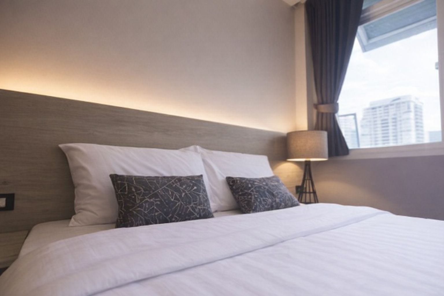 Hotellrum med dubbelsäng, sänggavel med belysning och fönster med stadsvy.