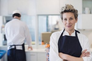 En leende kock med kort hår och mörkblått förkläde framför en kollega i ett storkök.