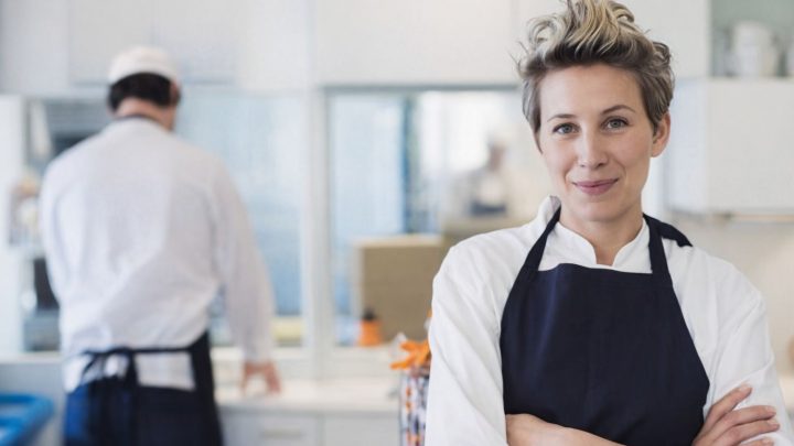 En leende kock med kort hår och mörkblått förkläde framför en kollega i ett storkök.