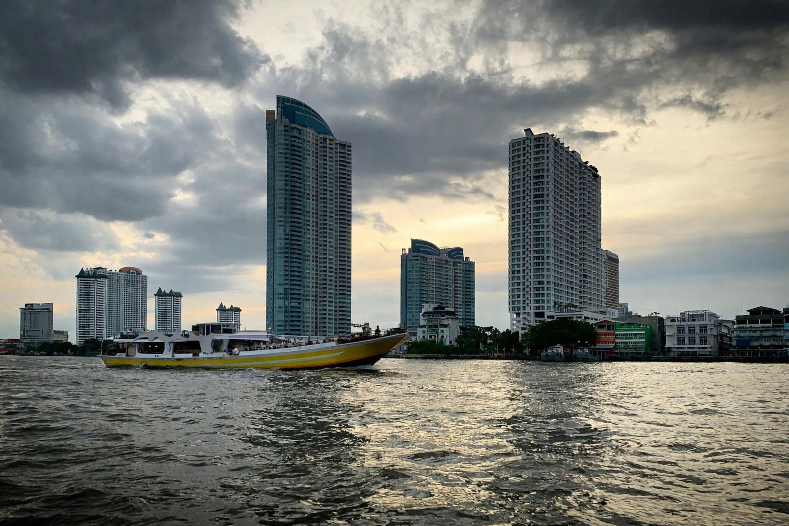 Båt på floden vid höghus under molnig himmel.
